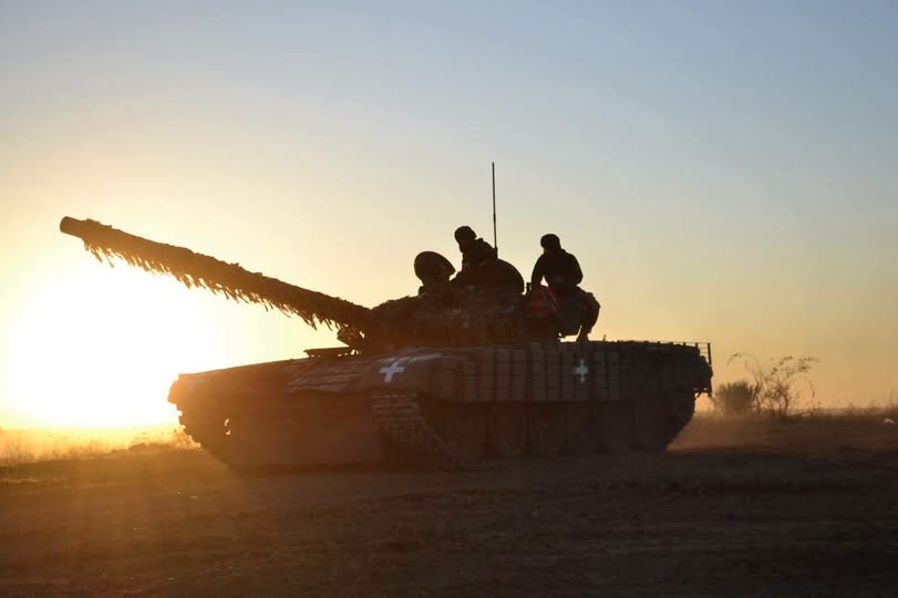 Силует українського танка із замаскованим стволом та екіпажем на броні рухається полем на тлі яскравого вечірнього або ранкового сонця, що заходить за горизонт