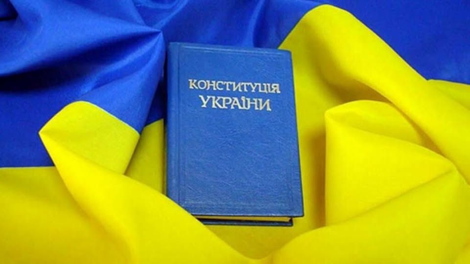 На зображенні представлена синя книга з тисненим золотим написом «КОНСТИТУЦІЯ УКРАЇНИ», яка лежить у центрі на об'ємних складках синьо-жовтого державного прапора.
