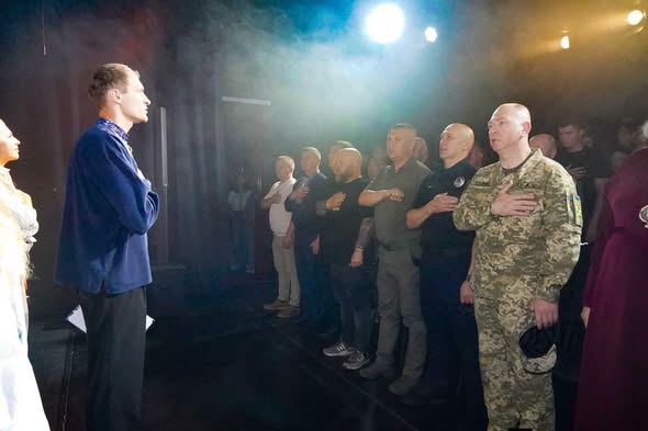 На світлині зображено чоловіка в синій вишиванці на сцені, який разом із присутніми в залі військовими, поліцейськими та цивільними тримає руку на серці під час виконання державного гімну в освітленому прожекторами та димом приміщенні.