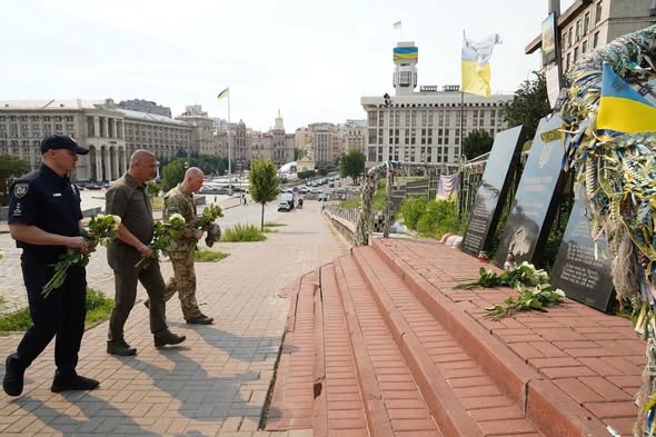 На світлині зображено трьох чоловіків в уніформах (поліцейського, військового та службовця), які з білими квітами в руках підходять Алеєю Героїв Небесної Сотні у Києві до меморіальних плит із портретами загиблих для вшанування їхньої пам'яті.