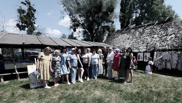 Група людей стоїть у ряд на траві біля дерев'яних торговельних яток та традиційної будівлі зі стріхою під час ярмарку просто неба в оточенні дерев.