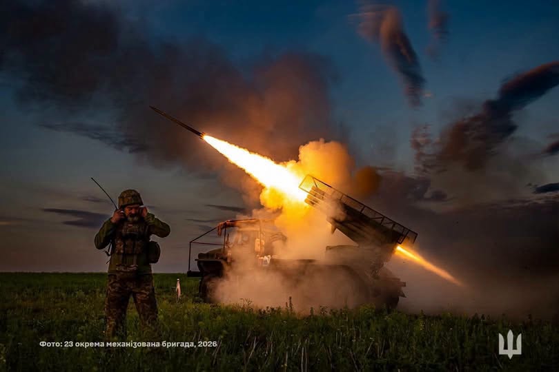 На зображенні показано вечірній запуск ракети з української реактивної системи залпового вогню, де військовослужбовець на передньому плані закриває вуха руками від гучного звуку пострілу