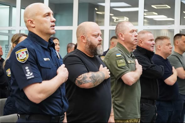 Ряд чоловіків у військовій, поліцейській та цивільній формі стоять у приміщенні та тримають праву руку на серці під час урочистої події або виконання гімну.