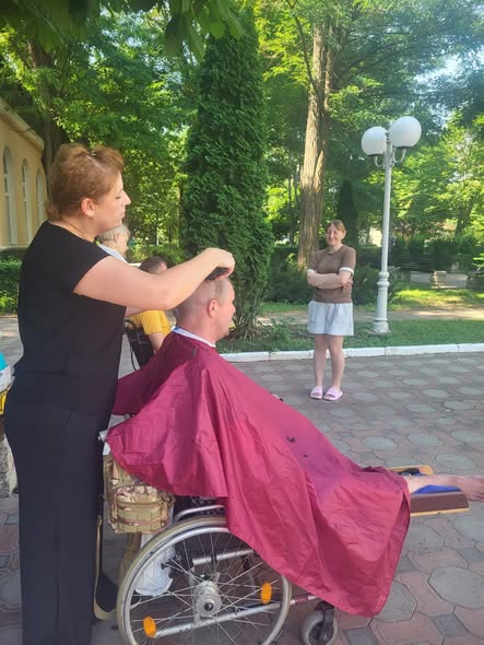 На світлині просто неба у парку жінка-перукар стриже чоловіка, який сидить у колісному кріслі, загорнутий у бордову накидку-пеньюар, тоді як на задньому плані за ними спостерігає інша усміхнена жінка.