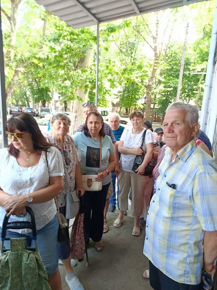 Група людей середнього та літнього віку стоїть у черзі під навісом будівлі на тлі зелених дерев вулиці.