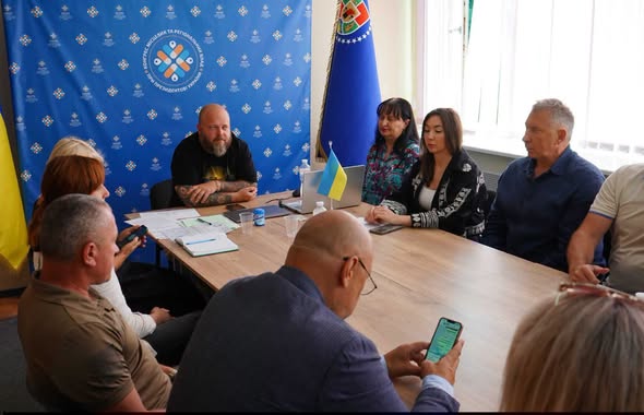 На фотографії зображено робочу зустріч групи людей за довгим дерев'яним столом у кабінеті на тлі синього банера з логотипом Конгресу місцевих та регіональних влад і прапорів, де учасники обговорюють документи, використовують ноутбук та мобільний телефон.