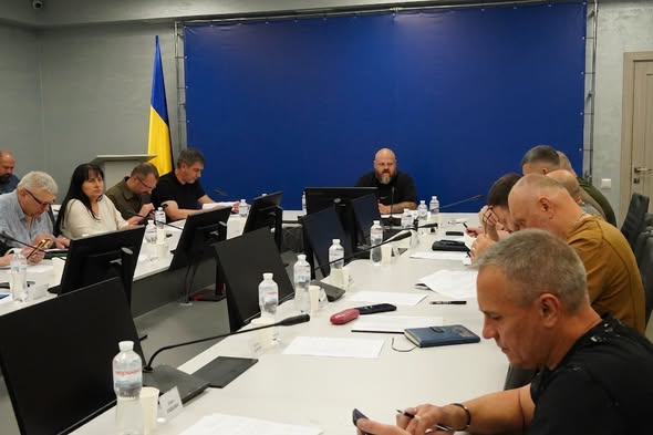 Офіційне засідання групи людей за великим П-подібним столом із моніторами, де в центрі на тлі синього банера сидить лисий бородатий чоловік, а ліворуч у кутку стоїть прапор України.
