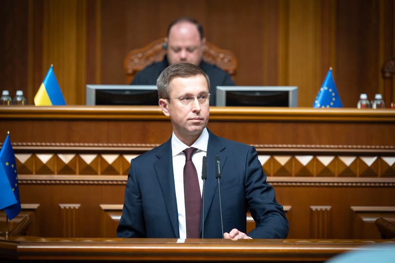 Світлина із сесійної зали Верховної Ради України, де за трибуною з мікрофонами стоїть чоловік у темно-синьому костюмі та окулярах, а на задньому плані за дерев'яною президією з прапорами України та ЄС сидить інший чоловік.