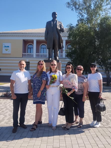 Група з шести людей у літньому повсякденному одязі, деякі з яких тримають квіти, стоїть на вимощеній площі та позує для спільного фото перед високим бронзовим пам'ятником чоловікові у костюмі на тлі двоповерхової адміністративної будівлі з колонами.