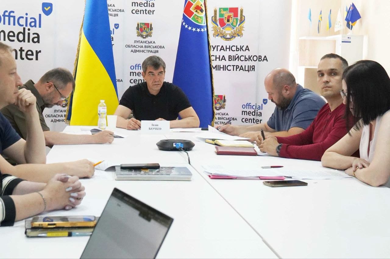 У світлому залі за великим білим столом із паперами та гаджетами проходять робочі збори, де в центрі на тлі прапорів та символіки Луганської обласної військової адміністрації сидить спікер із табличкою «Іван БОЧКО», а з обох боків від нього розмістилися інші учасники та учасниці зустрічі.
