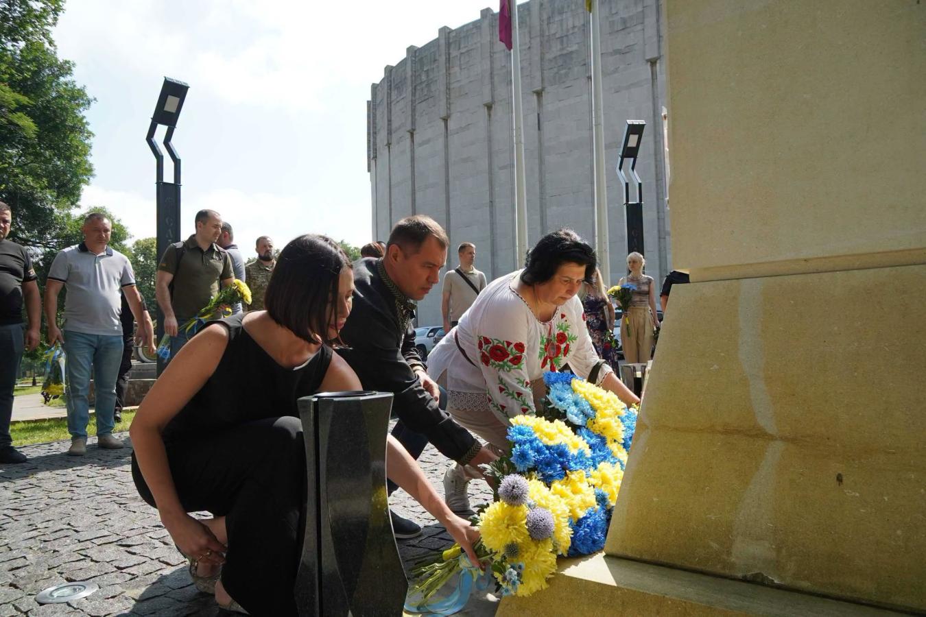 Люди, серед яких жінка у вишиванці, присіли біля підніжжя меморіалу на брущатці, щоб покласти великі синьо-жовті букети квітів під час офіційної церемонії вшанування пам'яті на тлі круглої сірої будівлі та інших учасників заходу.