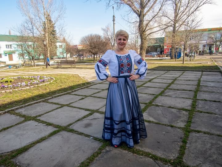 На світлині посеред скверу стоїть усміхнена жінка у вишиванці та довгій блакитній спідниці з прозорим верхнім шаром, тримаючи руки на талії