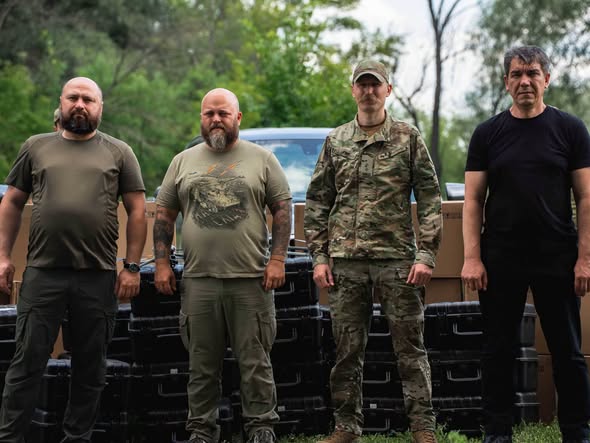 На тлі зелених дерев та коробок стоять четверо чоловіків у темному одязі та військовій камуфляжній формі, троє з яких дивляться прямо в кадр, а один зліва злегка повернув голову.