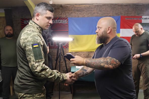 У приміщенні на тлі українського прапора та військової символіки татуйований чоловік у темній футболці тисне руку військовослужбовцю у камуфляжній формі та передає йому смартфон, поки двоє інших чоловіків спостерігають за процесом на задньому плані.