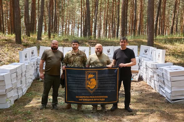 У сосновому лісі на тлі чисельних стосів білих картонних коробок стоять четверо чоловіків у військовому та цивільному одязі, які тримають перед собою великий чорний прапор із символікою та написом «60 ОКРЕМА МЕХАНІЗОВАНА ІНГУЛЕЦЬКА БРИГАДА».