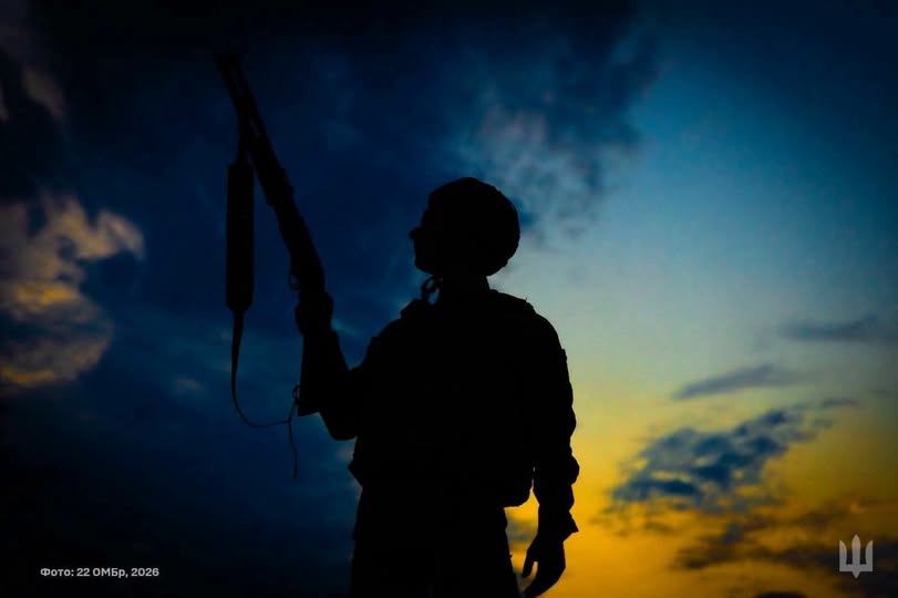 На фотографії зображено темний силует військовослужбовця в екіпіруванні, який тримає зброю, спрямовану вгору, на тлі вечірнього або ранкового неба з контрастними синіми та жовто-гарячими хмарами.