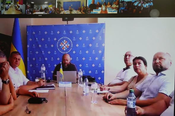 Скриншот робочої онлайн-наради, де на великому екрані видно учасників за столом у залі засідань на тлі синього банера з емблемою, а у верхній частині кадру відображаються маленькі віконця інших підключених учасників відеоконференції.