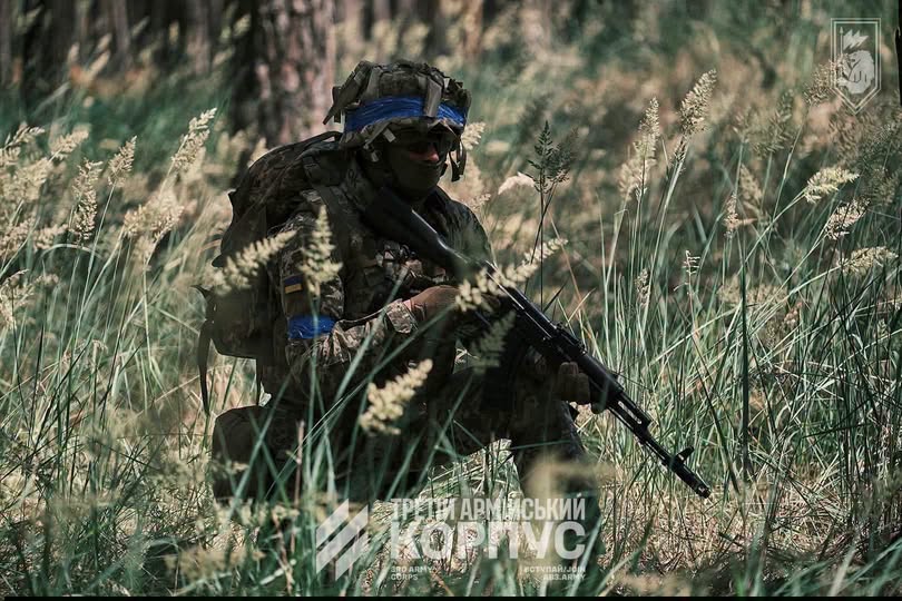 На світлині зображено українського військовослужбовця у повному бойовому екіпіруванні з автоматом у руках, який присів серед високої трави у лісистій місцевості, а знизу розміщено текстовий логотип «ТРЕТІЙ АРМІЙСЬКИЙ КОРПУС».