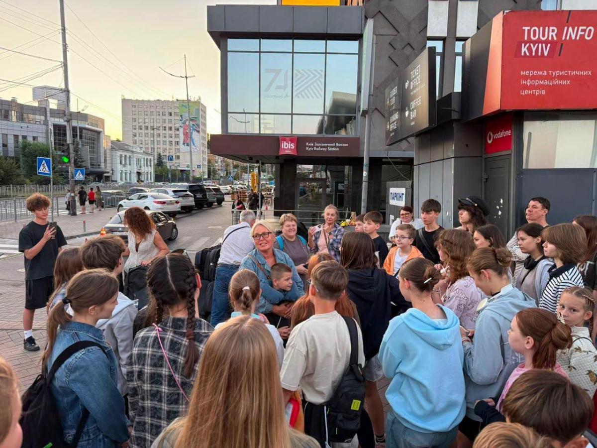 Великі групи дітей та дорослих зібралися на вулиці біля залізничного вокзалу в Києві поруч із будівлею готелю «Ibis» та туристичним інфоцентром, де жінка в окулярах щось розповідає дітям, які стоять півколом до неї.
