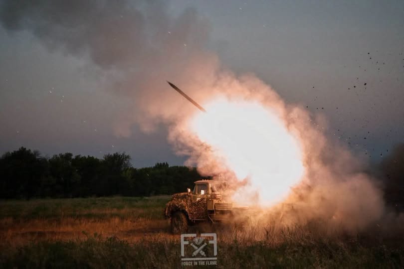 На зображенні у вечірніх сутінках зафіксовано момент пострілу з військової вантажівки, яка випускає ракету в небо серед яскравого спалаху вогню, густих клубів диму та розлітання землі на тлі силуетів дерев.