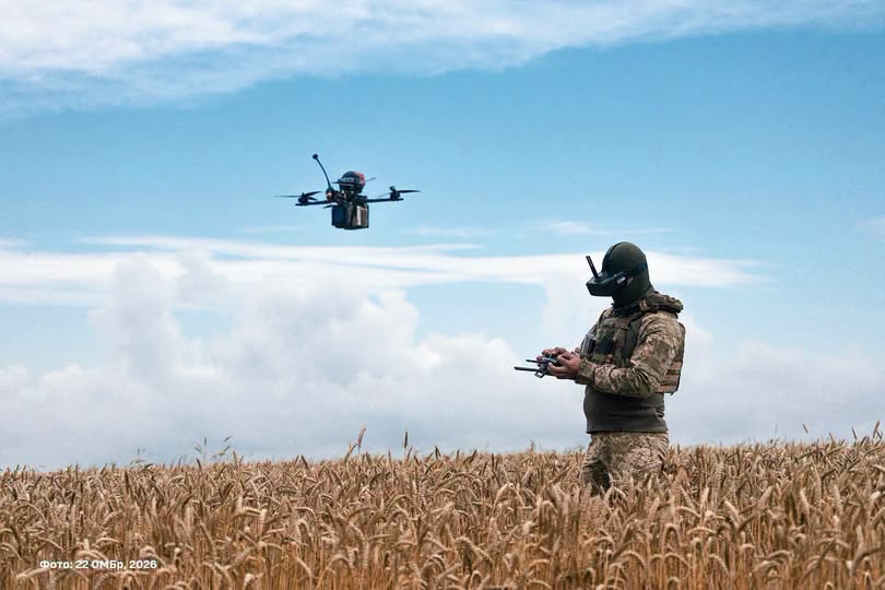 На фото зображено військового у камуфляжній формі та FPV-окулярах, який стоїть серед стиглого пшеничного поля під хмарним синім знаком та керує пультом безпілотником, що летить у повітрі.