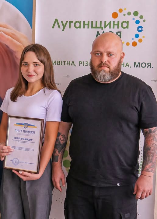 На фото зображено молоду жінку у білій футболці, яка тримає в руках рамку з листом подяки, та чоловіка міцної статури з бородою й татуюваннями на руках у чорній футболці, які стоять поруч на тлі банера з написом «Луганщина».
