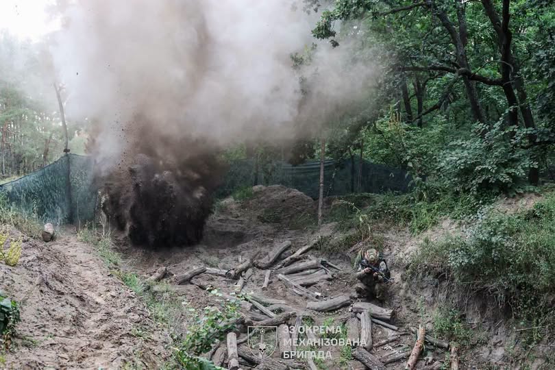 На зображенні зафіксовано момент потужного вибуху із викидом густого темного диму та землі у лісистій місцевості, де на передньому плані в окопі з колод сидить український військовослужбовець із зброєю, а внизу по центру розміщено емблему та напис «33 окрема механізована бригада».