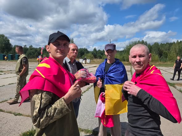 Група чоловіків на відкритому майданчику, деякі з яких огорнуті прапорами (синьо-жовтим та малиновими), стоять на тлі дерев і хмарного неба, а чоловік на передньому плані ліворуч показує жест «великий палець вгору».