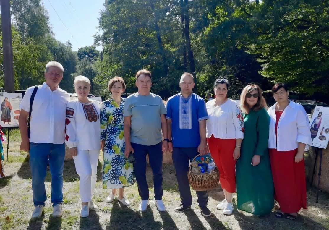 На тлі густих зелених дерев сонячного дня зображено групу з восьми людей, які вишикувалися в ряд у повсякденному та традиційному вишиваному одязі, причому один із чоловіків у центрі тримає плетений кошик із продуктами, а зліва та справа на задньому плані видніються інформаційні стенди.