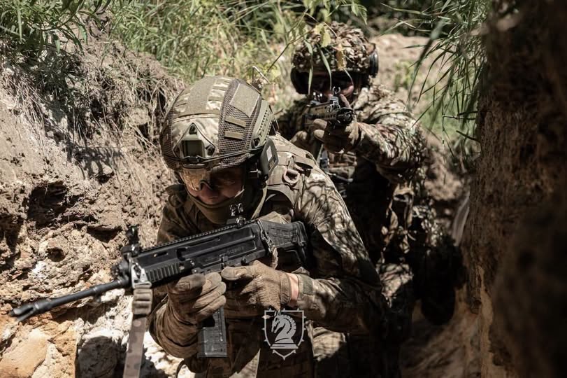 Двоє військовослужбовців у камуфляжній формі та захисному спорядженні зі зброєю в руках просуваються всередині вузького земляного окопу.
