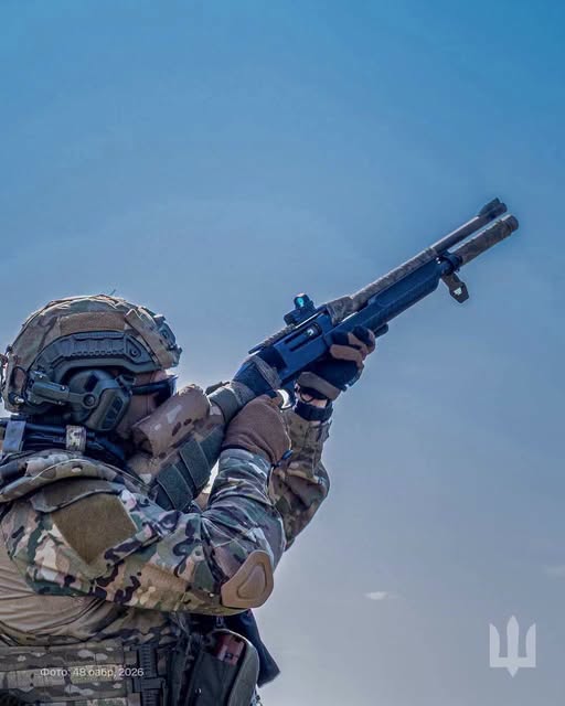 На зображенні показано військовослужбовця у камуфляжній формі, шоломі та тактичних навушниках, який цілиться вгору на тлі чистого блакитного неба з напівавтоматичної рушниці, обладнаної коліматорним прицілом.