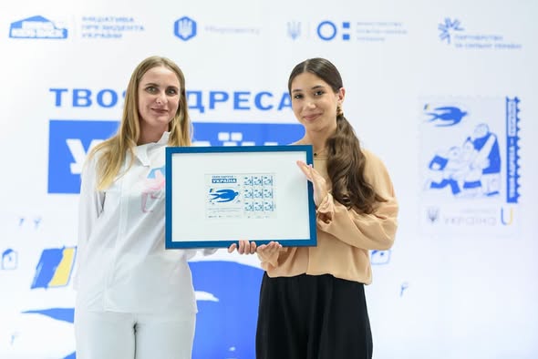 Дві усміхнені молоді жінки стоять поруч на тлі білого банера з синіми написами та разом тримають у руках прямокутну синю рамку із зображенням аркуша поштових марок.