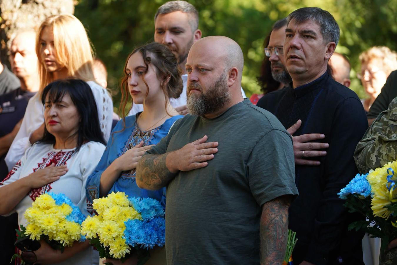Група людей у вишиванках та цивільному одязі зосереджено стоїть на вулиці, притиснувши праву руку до серця під час урочистої події, тримаючи в руках пишні синьо-жовті букети хризантем.