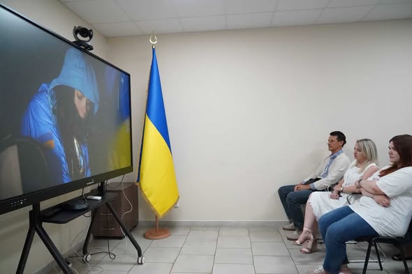 У світлій кімнаті з державним прапором України троє людей сидять на стільцях і дивляться на великий монітор, де на екрані транслюється зображення звільненої з полону жінки, загорнутої в синьо-жовтий стяг.