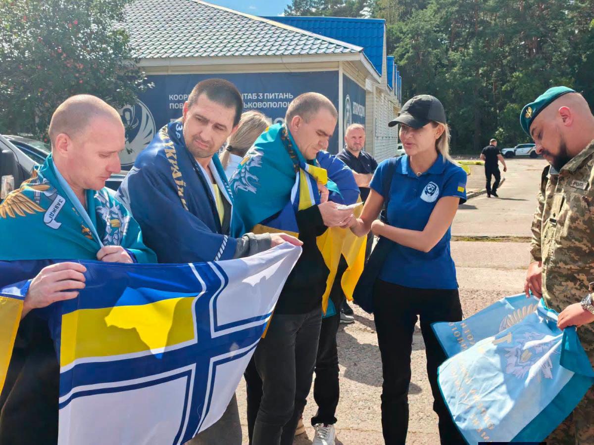 На зображенні група людей просто неба, серед яких звільнені з полону українські військовослужбовці, загорнуті в державні прапори та прапори різних родів військ, спілкуються з представницею Координаційного штабу та військовим у камуфляжі.
