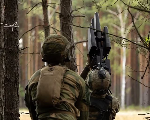 Двоє військовослужбовців у захисних шоломах та камуфляжній формі зафіксовані зі спини серед соснового лісу, де один із них спрямовує вгору спрямовану антену портативного комплексу радіоелектронної боротьби — так звану «антидронову рушницю».