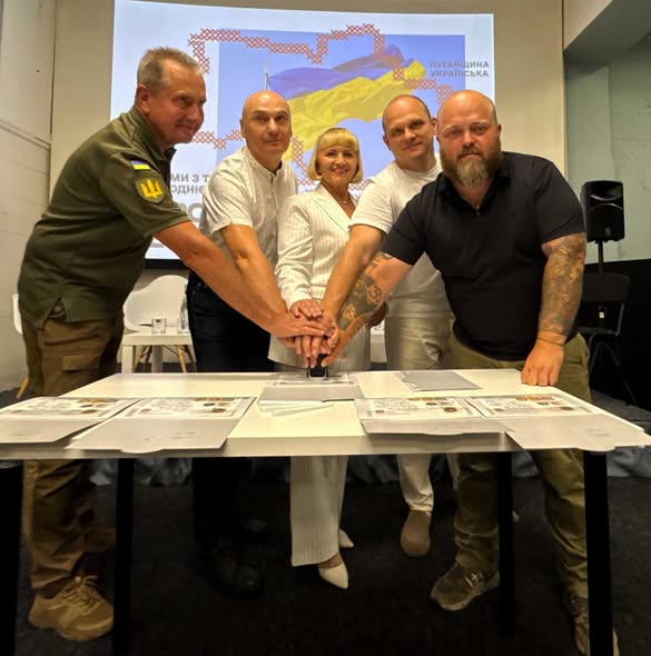 П'ять людей, серед яких чоловік у військовій формі, жінка у світлому брючному костюмі та троє чоловіків у цивільному одязі, склали руки разом для символічного спецпогашення поштових марок за столом на тлі екрана з написом «Луганщина українська».