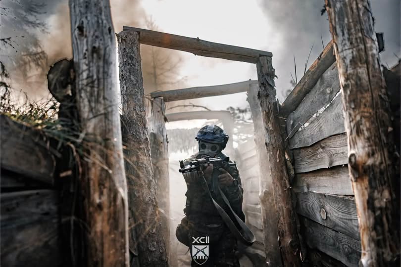 На світлині крізь задимлений дерев'яний окоп просувається військовослужбовець у касці та протигазі, який цілиться вперед з автоматичної зброї.