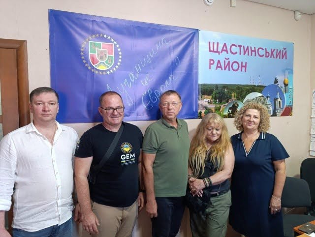 Група з трьох чоловіків та двох жінок стоїть у приміщенні на тлі великого синього прапора з гербом Луганської області та плаката з написом «Щастинський район».