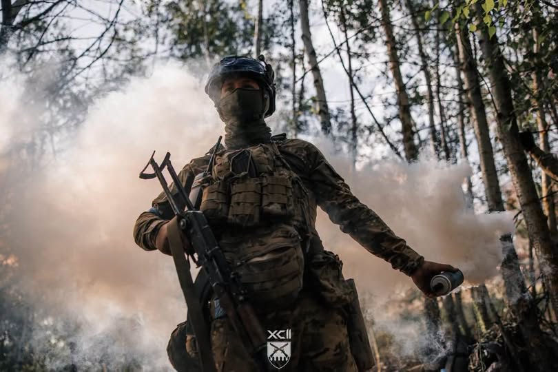 На світлині в лісовій місцевості військовослужбовець у повному бойовому спорядженні, касці та балаклаві тримає автомат в одній руці та відводить убік іншу руку з димовою шашкою, від якої за його спиною розходиться густа хмара білого диму.