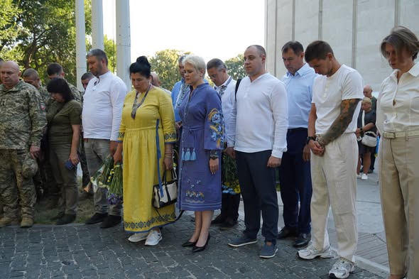 Група людей у цивільному одязі, вишиванках та військовій формі зосереджено стоїть із нахиленими головами на брукованій площі під час хвилини мовчання чи офіційної церемонії пам'яті.