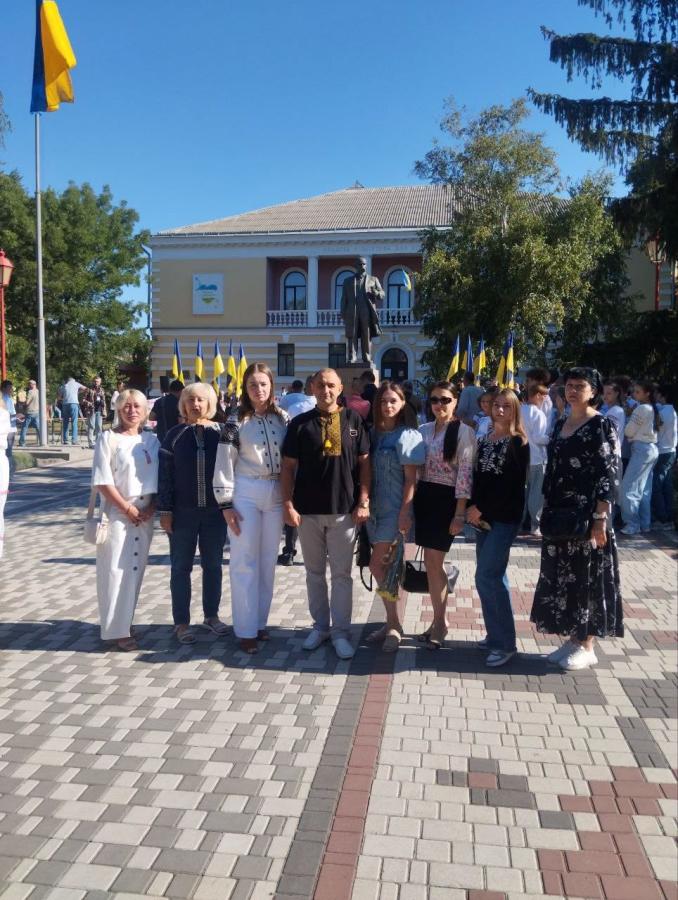 Група людей у вишиванках та літньому одязі стоїть на брукованій площі міста для спільного фото на тлі пам'ятника Тарасові Шевченку, багатьох синьо-жовтих прапорів та двоповерхової будівлі під ясним синім небом.