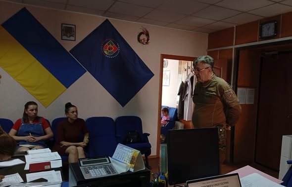 На світлині в робочому кабінеті сивий чоловік у військовій формі стоїть і спілкується з двома жінками, які сидять у синіх кріслах на тлі стіни, де розвішані Державний Прапор України та великий синій прапор з емблемою.