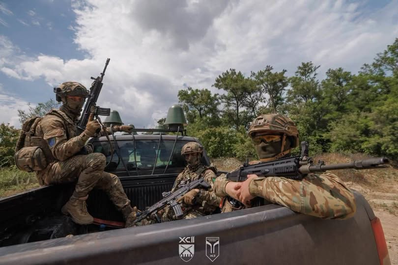 Три військові у камуфляжній формі та зі зброєю в руках перебувають у кузові пікапа на тлі дерев та хмарного неба.