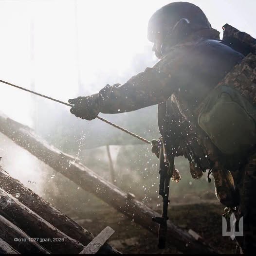 На зображенні військовослужбовець у касці та спорядженні тримає натягнутий трос на тлі дерев'яних колод бліндажа, а яскраве контрве світло підсвічує дрібні бризки та дим навколо