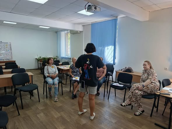У світлій навчальній аудиторії жінка в чорній футболці з малюнком стоїть спиною до глядача по центру кімнати і спілкується з чотирма іншими жінками, які сидять півколом на чорних стільцях біля парт