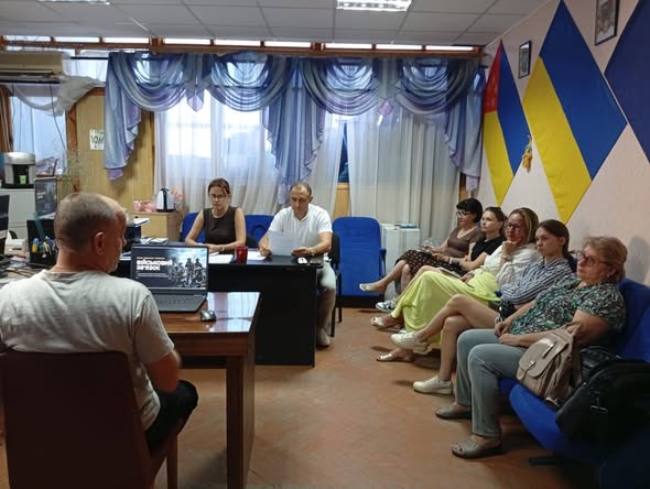 У кабінеті з прапором України на стіні та синіми шторами на вікнах чоловік за столом із ноутбуком проводить зустріч для групи людей, які сидять на стільцях та диванах уздовж кімнати