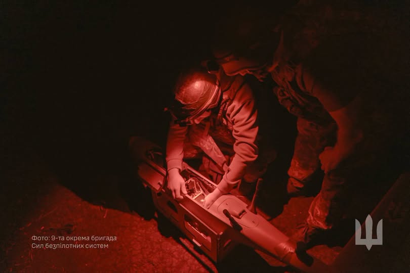 На зображенні в умовах червоного нічного освітлення двоє військових налаштовують безпілотний літальний апарат, а в нижньому кутку розміщено напис із назвою підрозділу та герб України.