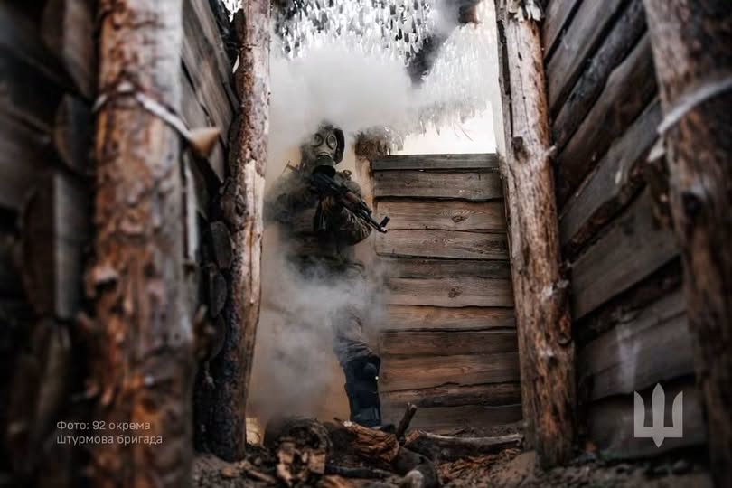 На зображенні військовослужбовець у протигазі та повному бойовому екіпіруванні стоїть з автоматом у руках всередині дерев'яного бліндажа або окопу, оповитого густим білим димом.
