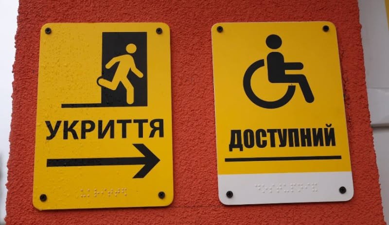 На яскраво-помаранчевій текстурній стіні закріплено дві жовті інформаційні таблички: ліва містить піктограму людини, що біжить до дверей, слово «УКРИТТЯ» та стрілку праворуч, а права — міжнародний символ доступності для людей на кріслах колісних та напис «ДОСТУПНИЙ», причому обидві таблички мають знизу дублювання шрифтом Брайля.
